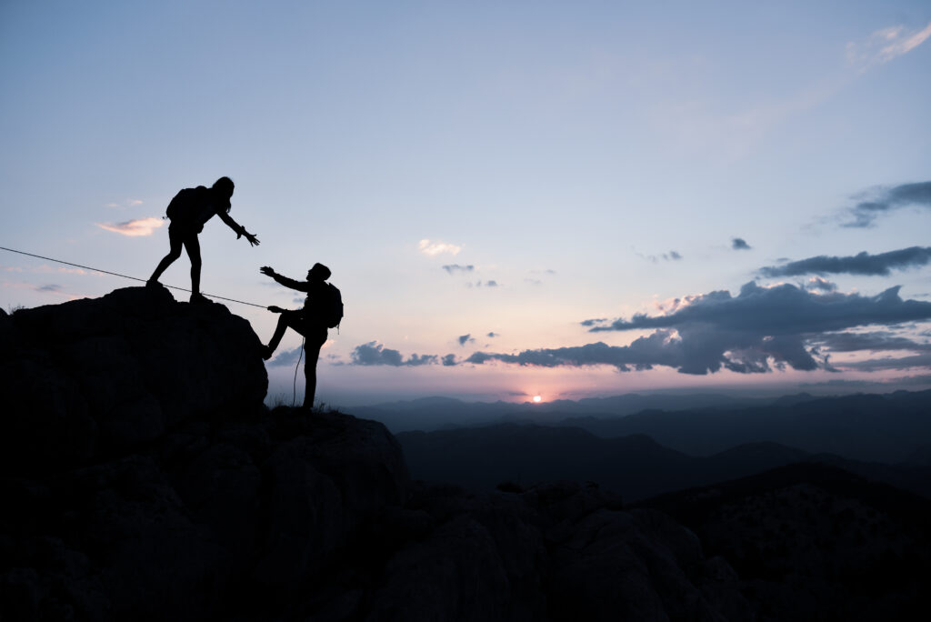 "Deux randonneurs s’entraident lors d’une ascension au coucher du soleil, illustrant l’entraide, le dépassement de soi et l’harmonie avec la nature